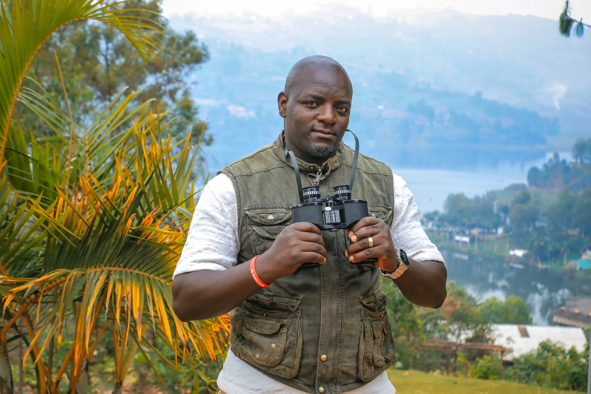 A man in a field waistcoat holding binoculars, with the lake and hills behind